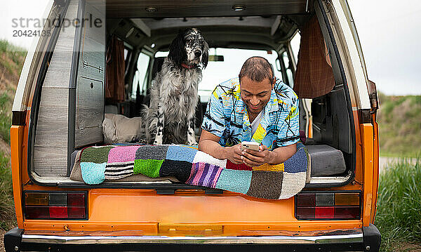 Happy ethnic man lying in van with dog and browsing smartphone