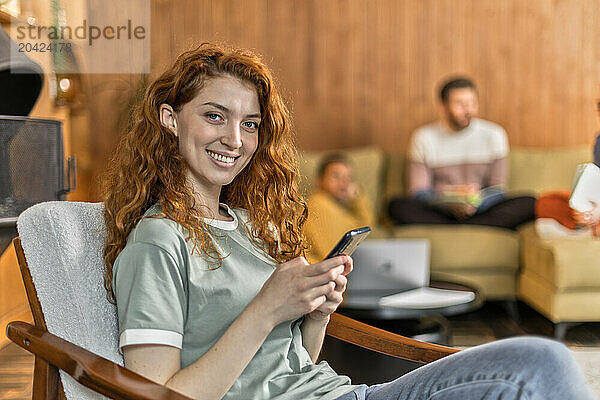 red haired young woman sit on armchair using mobile phone smiling