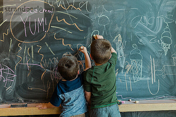 Two young brothers drawing colorful pictures on a large chalkboard.