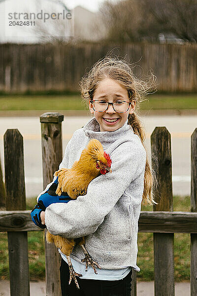 Tween girl in glasses and blond hair holds golden chicken in yard