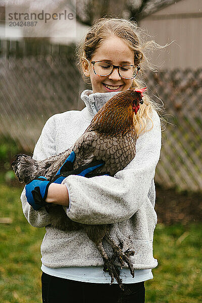 Tween blond girl in glasses smiles holding pet chicken