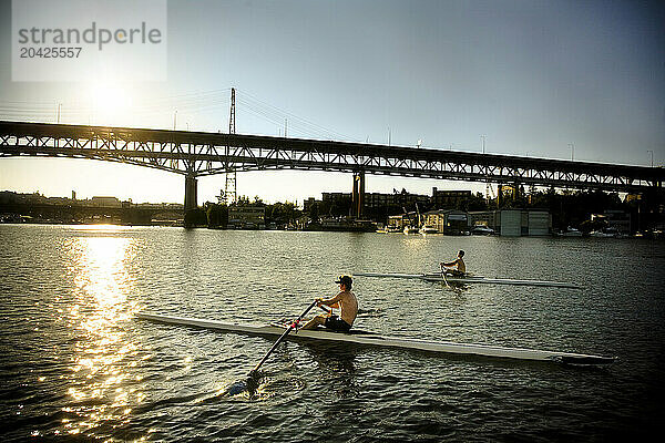 Two young men row single boats on a lake.