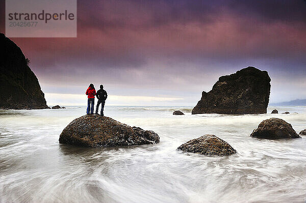 A man and a woman stand on a small rock surrounded by ocean water from the incoming tide  in Olympic National Park  Washington.