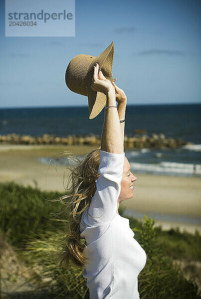 A woman raises her hat in the air and enjoys the sun and wind on the beach in Dennis  Massachusetts.