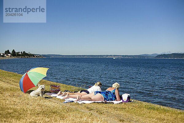 Two older women sunbathe with their dog under a red umbrella near Alki Beach  Seattle WA.