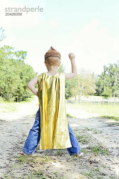 Child in superhero costume triumphantly raising fist outdoors.