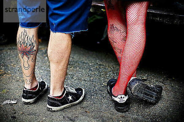 View of a man's tattooed legs and a woman's legs wearing red fishnet stockings standing on cement.