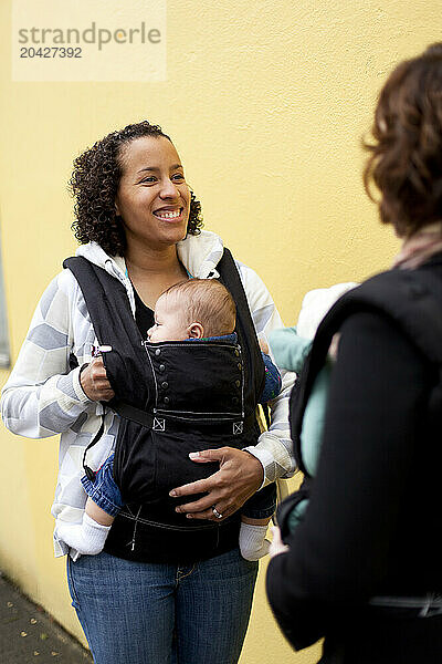 A smiling woman with a newborn baby in a chest carrier talks with a woman with a newborn baby also in a chest carrier in the city.