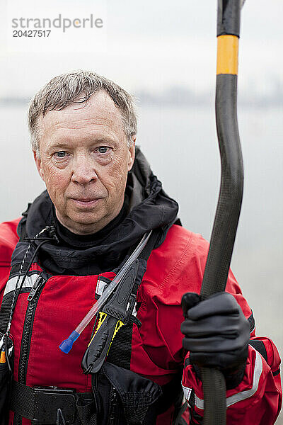 A portrait of a senior male kayaker in a red drysuit holding a carbon fiber paddle near the waters edge.