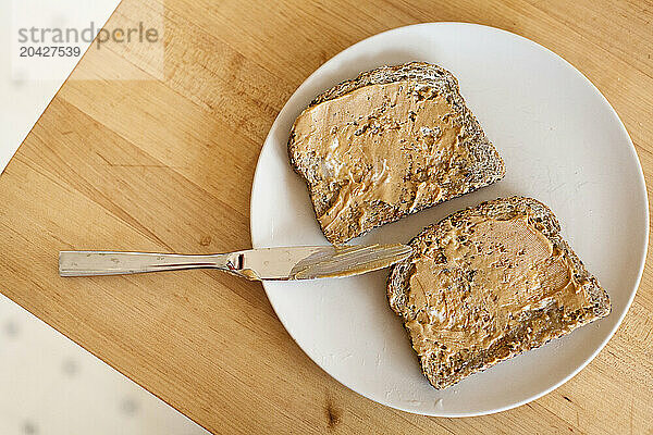 A top down view of peanut butter on toast on a plate with a knife on a kitchen table in a home.