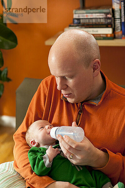 A father in orange feeds his newborn baby with a bottle while sitting on a couch in his home.