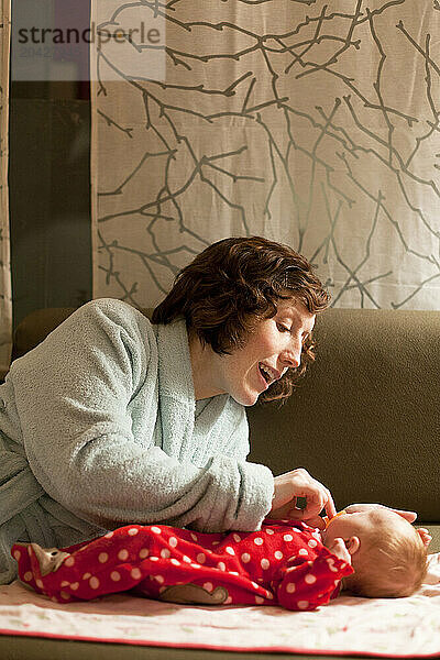 A mother in a blue bathrobe plays with her baby on a couch in her living room.