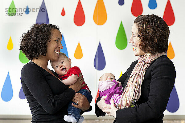 A smiling woman holds a newborn baby and talks with mom also holding a newborn baby at a park.