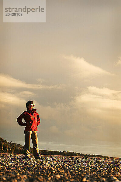 A young woman in an red jacket stands on a rocky beach at sunset in New Zealand.