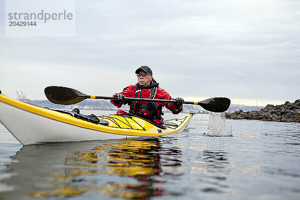 A senior male in his yellow kayak rows in front of the city skyline.