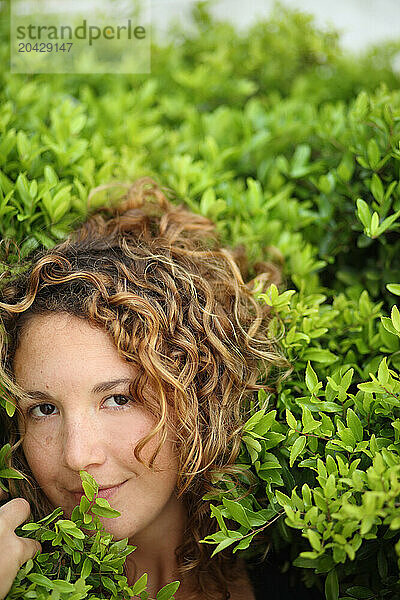 A curly haired woman looks coyly at the camera with her face surrounded by a bright green bush.