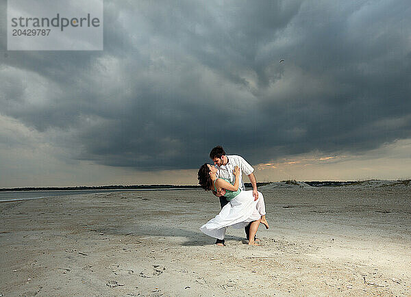 A man and a woman dance on the beach under a cloudy sky.