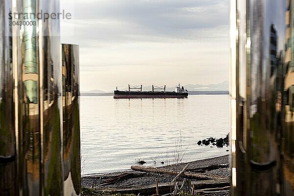 A large container ship is seen from the shoreline through an art installation.