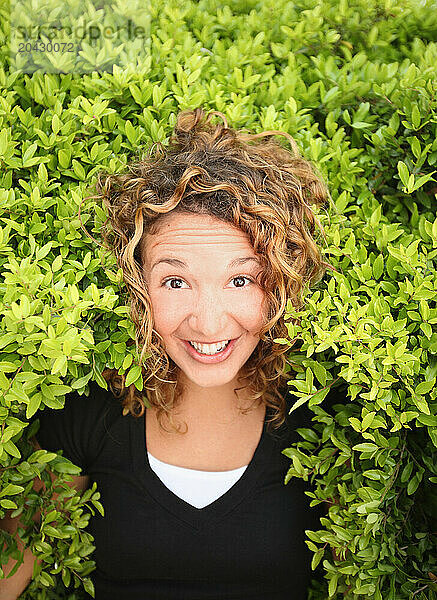 A young woman smiles at the camera as she peers out of a bright green bush.