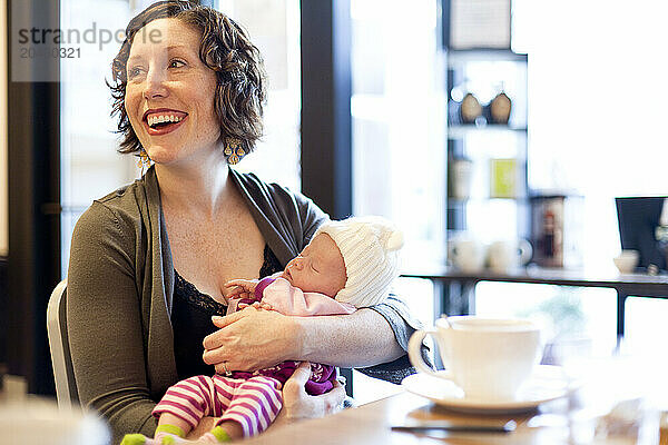 A smiling woman with red hair holds a sleeping newborn baby in a cafe.