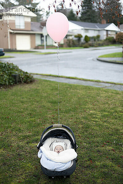 A newborn baby sits in a car seat on the grass alone  with a pink balloon floating above her.