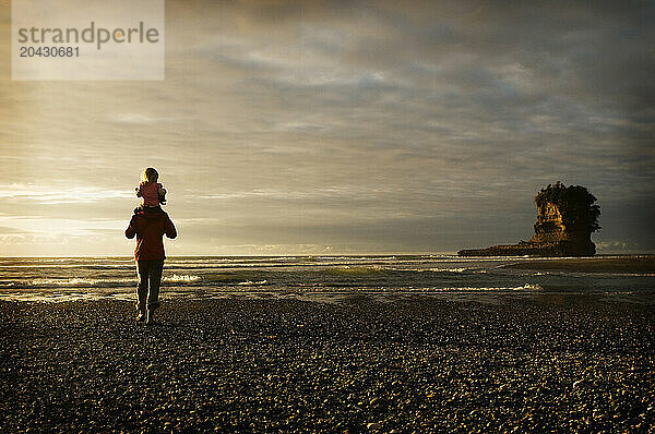 A young girl rides on her mothers shoulders while walking along a rocky beach at sunset in new Zealand.