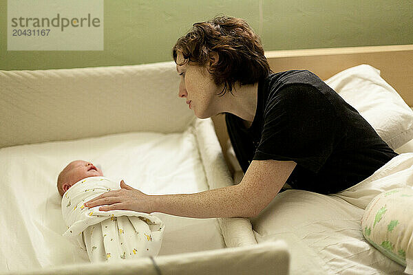 A woman soothes a swaddled crying baby in a co-sleeper crib next to her bed.