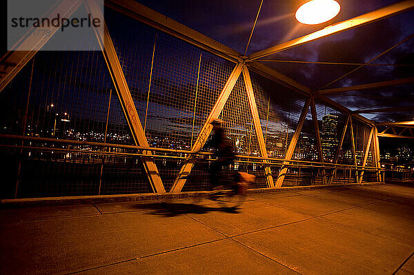 A bicyclist is blurred as he crosses a bridge in early evening with the skyline of Portland  Oregon lit up behind him on 2/21/2011