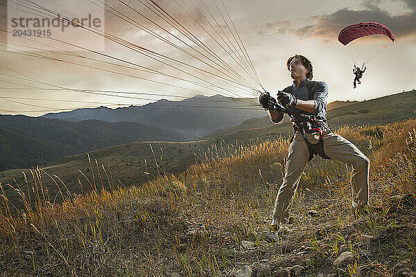 A speed flyer pulls his parasail in for a landing on a grassy hill at sunset.