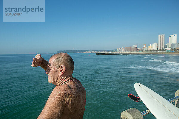 Senior man with surfboard watching for waves on pier near promenade on Golden Mile  Durban  South Africa