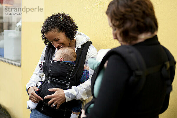 A smiling woman with a newborn baby in a chest carrier talks with a woman with a newborn baby also in a chest carrier in the city.