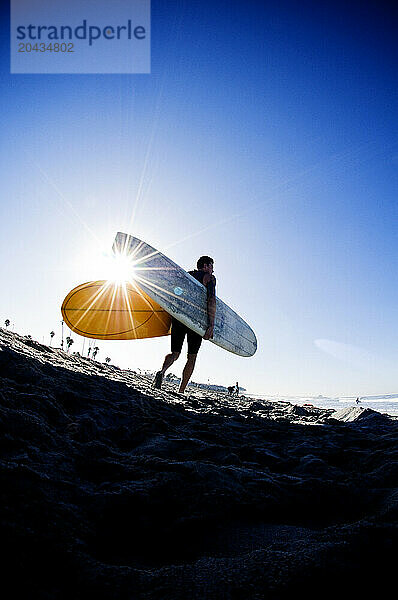 A young male surfer holding a surfboard with the sun shining behind him as he walks down a sandy beach.