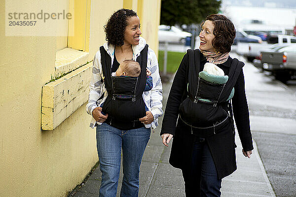 A smiling woman with a newborn baby in a chest carrier talks with a woman with a newborn baby also in a chest carrier in the city.