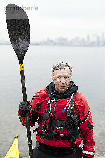 A portrait of a senior male kayaker in a red drysuit holding a carbon fiber paddle near the waters edge.