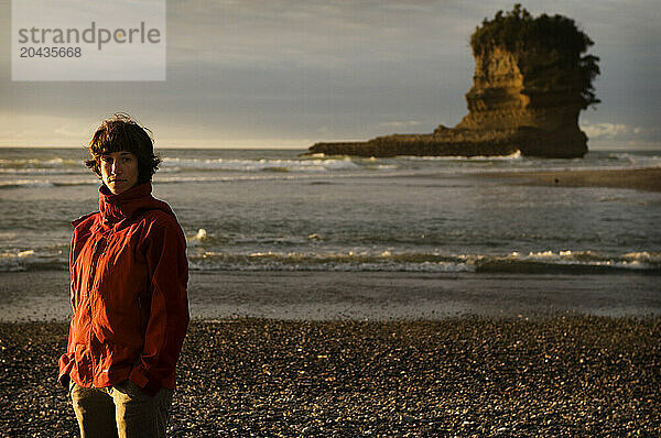 A young woman in an red jacket stands on a rocky beach at sunset in New Zealand.