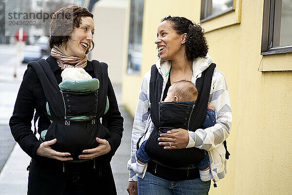 A smiling woman with a newborn baby in a chest carrier talks with a woman with a newborn baby also in a chest carrier in the city.