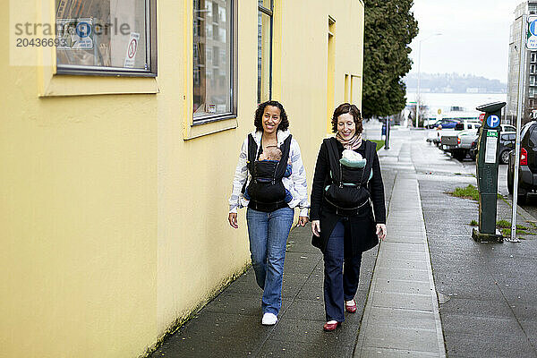 A smiling woman with a newborn baby in a chest carrier talks with a woman with a newborn baby also in a chest carrier in the city.