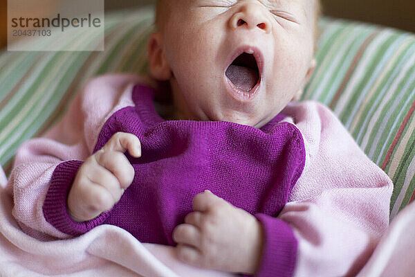 A newborn baby girl yawns while sitting on a couch in a home.