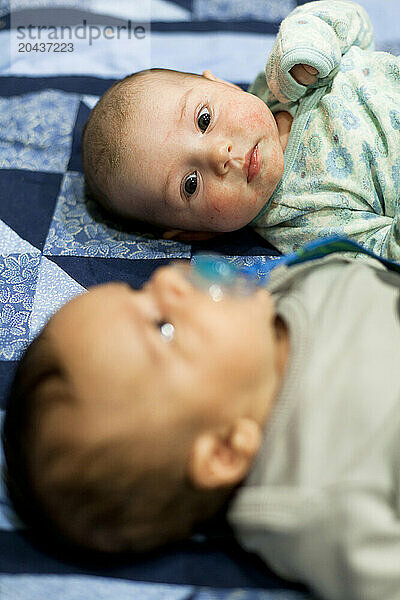 Two babies lie on a blue quilt in a home.