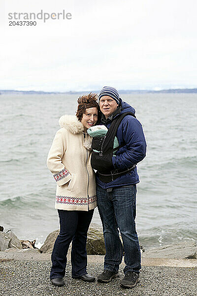 A father and mother pose for a picture with their newborn baby in a chest carrier on the beach during winter.