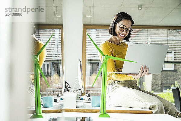 Smiling mature businesswoman using laptop sitting by wind turbine model on desk in office