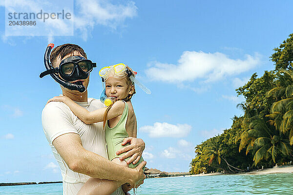 Father carrying daughter wearing snorkels and swimming goggles at beach