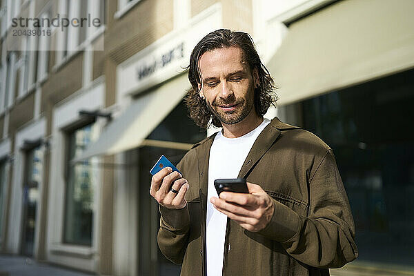 Smiling mature man using smart phone doing online shopping through credit card on sunny day