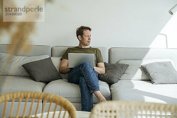 Thoughtful man with laptop working on sofa in living room