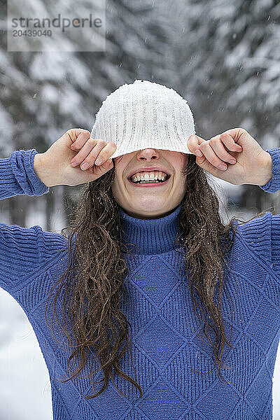 Smiling woman hiding face with knit hat in winter forest