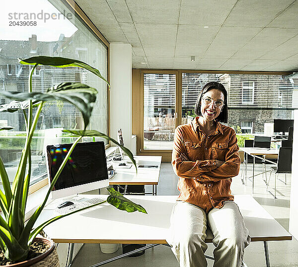 Happy mature businesswoman with arms crossed sitting on desk in office