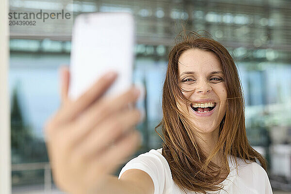 Laughing woman with brown hair taking selfie through smart phone at airport