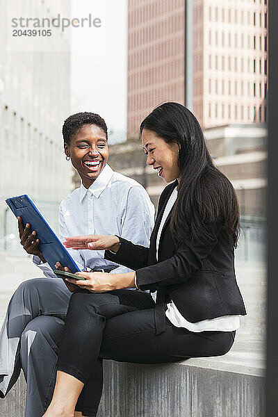 Cheerful businesswomen discussing over clipboard sitting at office park