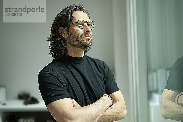 Thoughtful businessman standing with arms crossed looking out through window at office