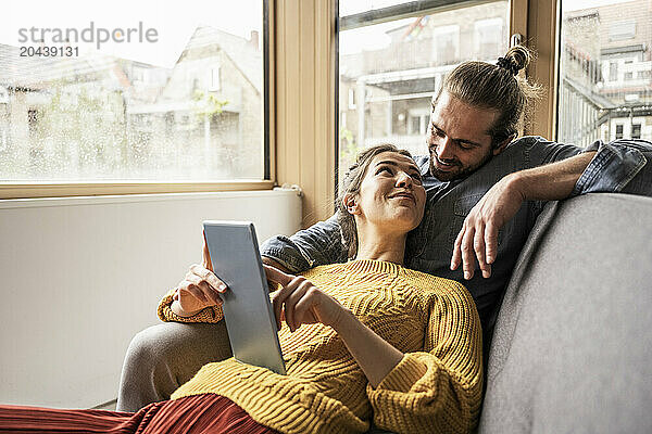 Smiling woman with tablet PC looking at boyfriend on sofa at home